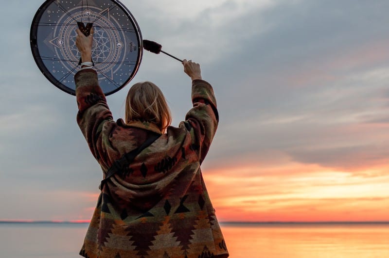 Femme qui fait du tambour chamanique sur une plage, devant un coucher de soleil