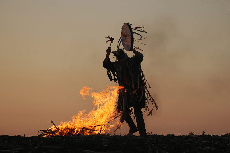 Un homme avec une tenue de chamane et un tambour danse autour d'un feu